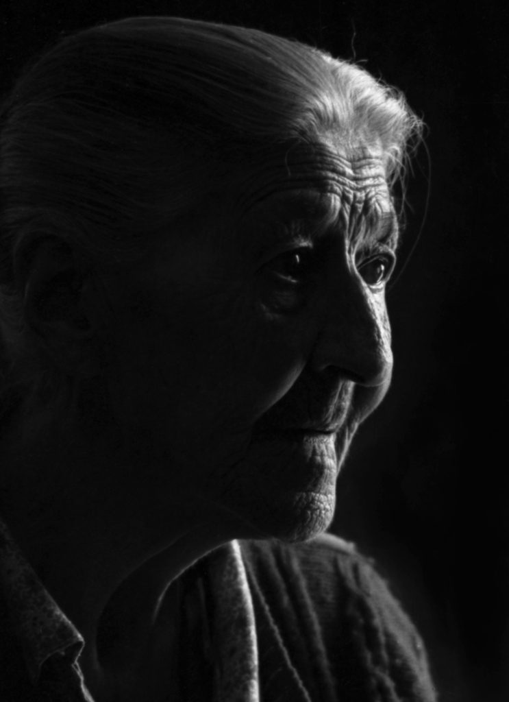 A black and white photo of an elderly woman mostly in subdued tones but illuminated by a rim of light picking her profile out of the darkness.