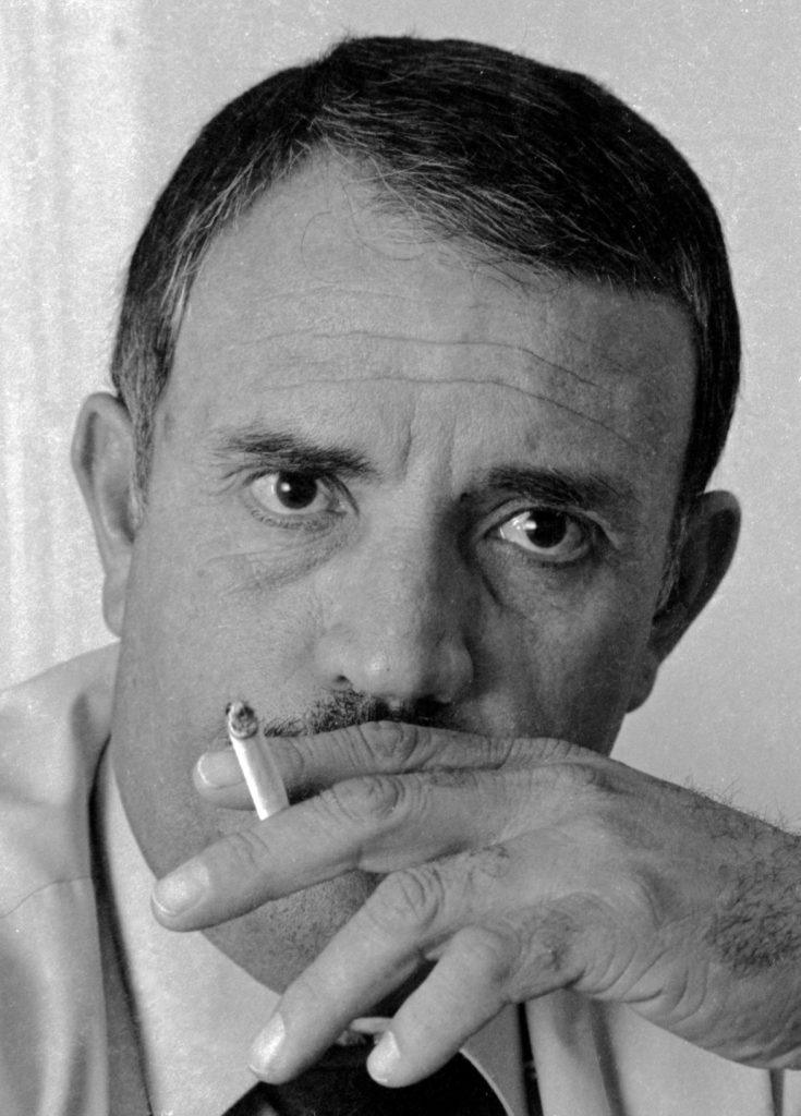 Image of a man with intent eyes. In front of his mouth and face is his hand holding a cigarette