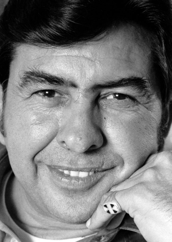 Close up of a man with a gentle smile leaning on his hand. The portrait is framed so his face almost fills the image.