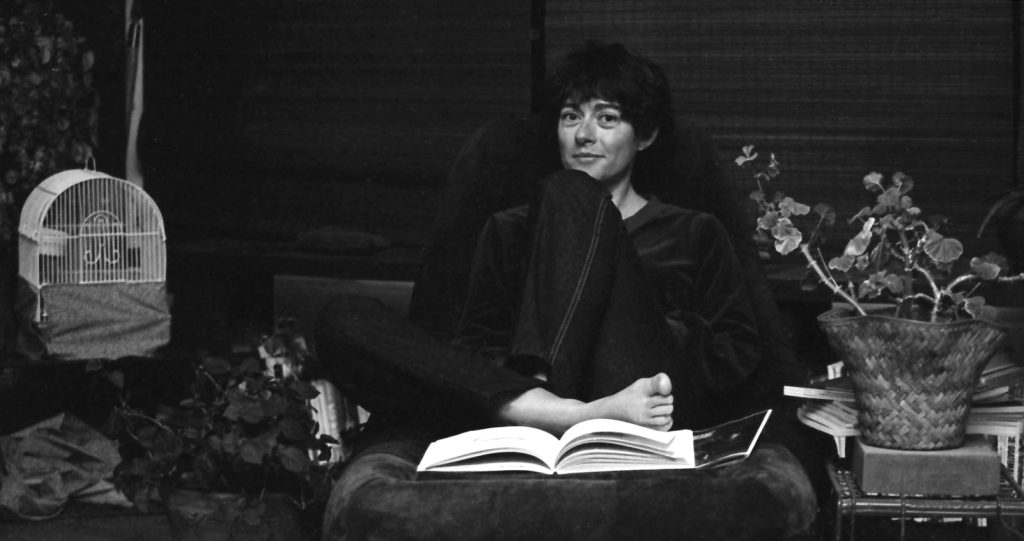 Full-body photo of an elegant woman sitting cross-legged on an armless stuffed chair with a birdcage to her right and a decorative plant to her left.