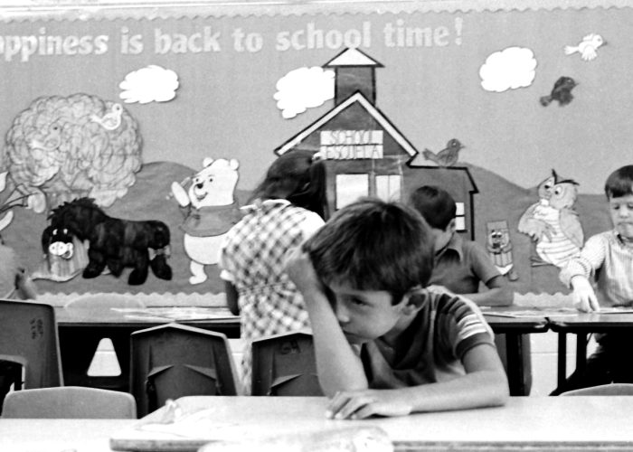 first-day-of-school-8-83 Young boy sitting at a table in a classroom, leaning forlornly on his hand. Behind him on the classroom wall are the words "Happiness is Back To School Time"