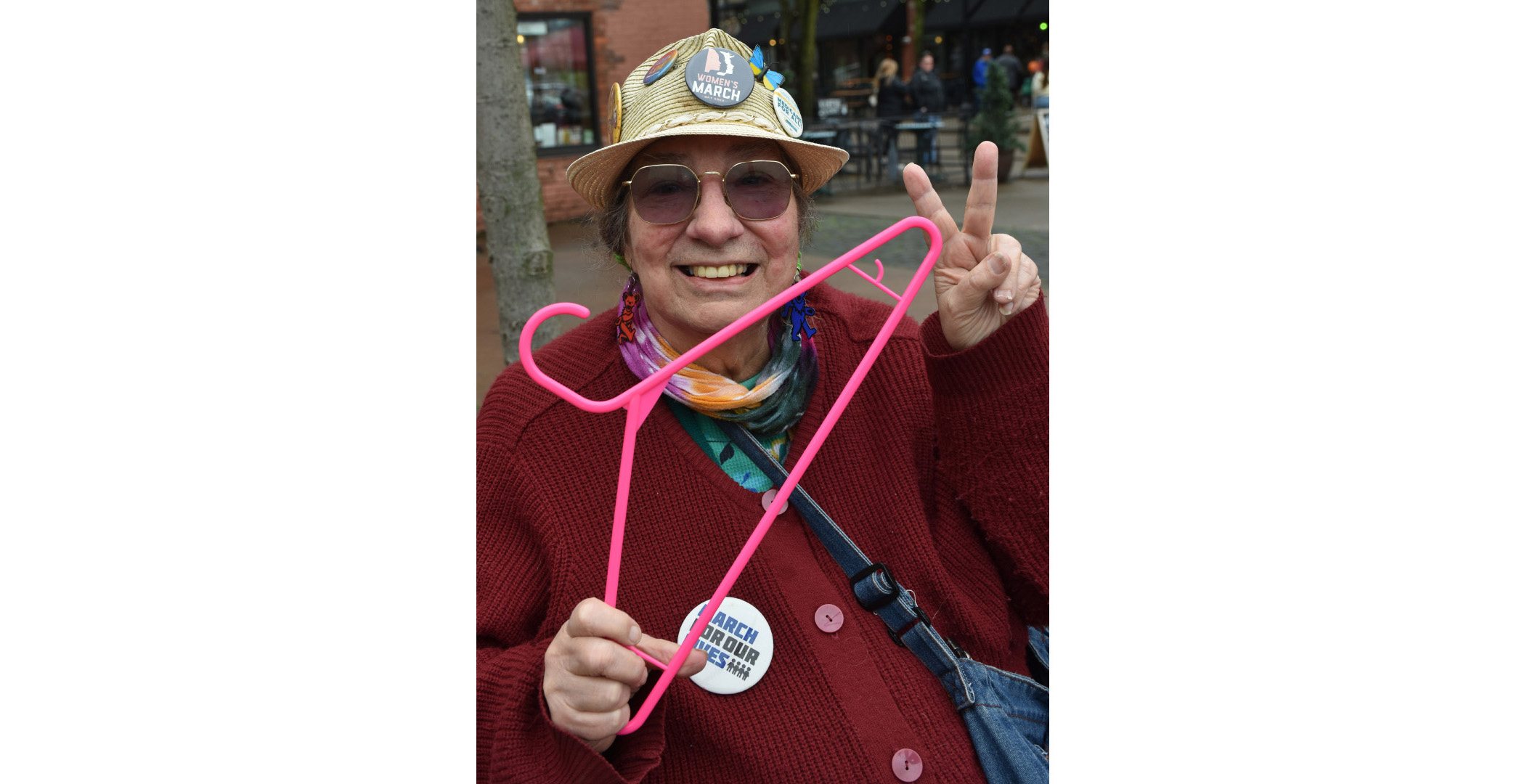 coathanger A smiling older woman in a red sweater holding a pink plastic coat hanger. She's wearing a hat full of buttons of protests she has attended over the years. She's wearing one more on her sweater that says "March For Our Lives."