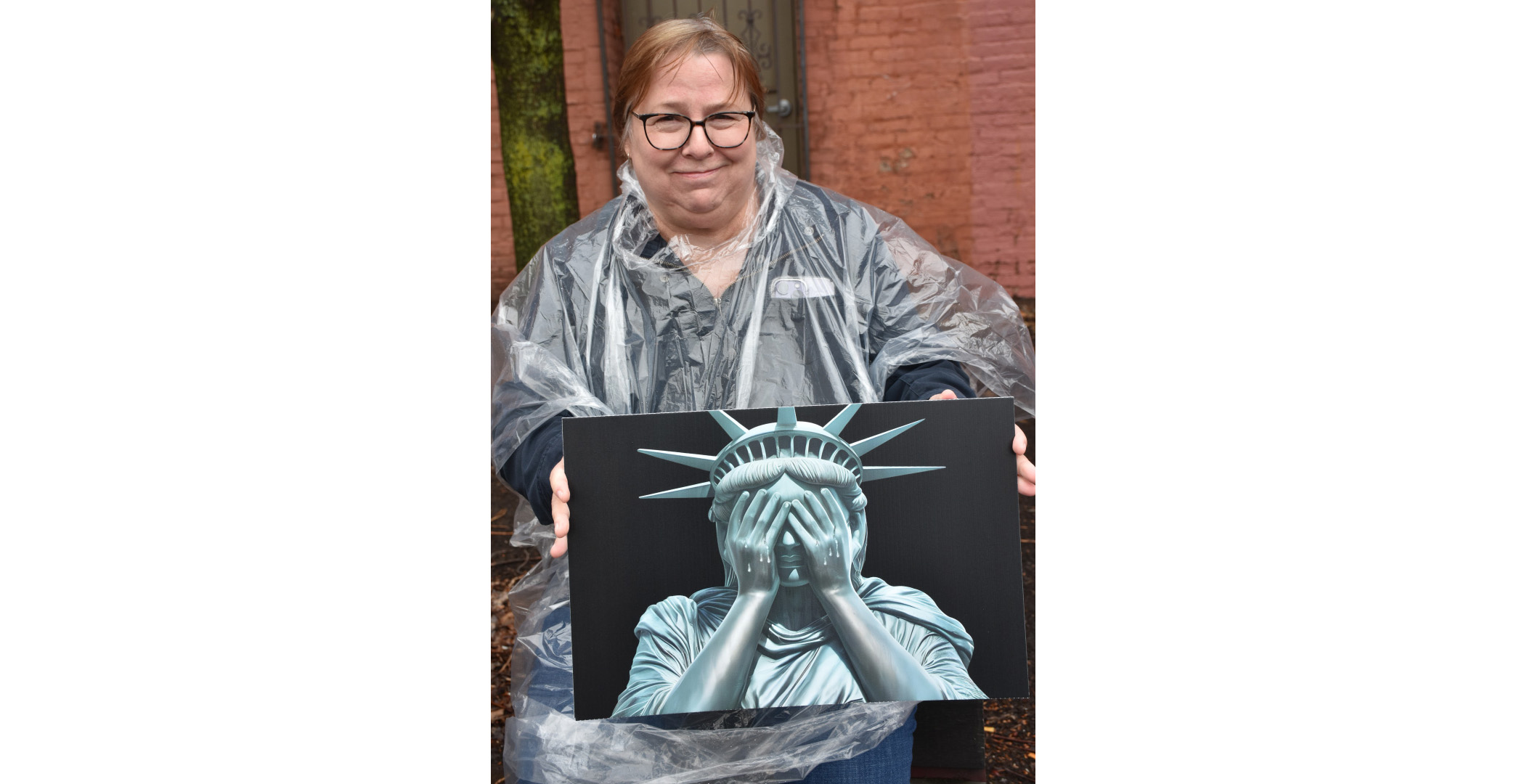 crying liberty A woman in a poncho holding a wordless sign with a realistic looking image of the head and shoulders and crown of the statue of liberty crying with her hands over her eyes A woman in a poncho holding a wordless sign with a realistic looking image of the head and shoulders and crown of the statue of liberty crying with her hands over her eyes
