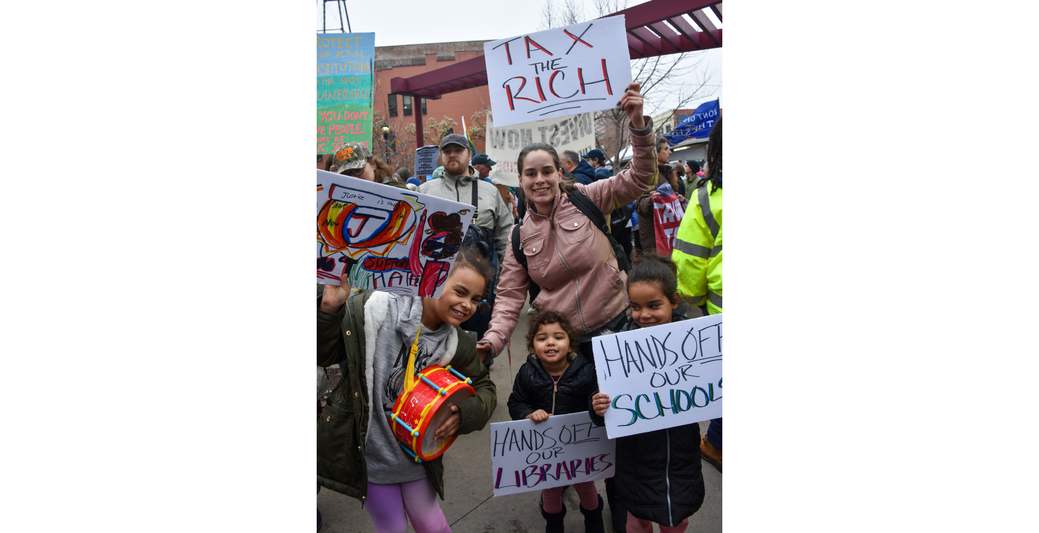 handsoff_protest4 Image shows a smiling woman with her young daughters at the Cleveland HandsOff protest on Market Square. The woman is holding a hand-made sign saying "Tax The Rich", the two younger kids have signs saying "Hands Off Our Schools," and Hands Off our Libraries." The older daughter's sign is hard to read in the photo but is colorfully decorated and she is holding a bright orange drum. Image shows a smiling woman with her young daughters at the Cleveland HandsOff protest on Market Square. The woman is holding a hand-made sign saying "Tax The Rich", the two younger kids have signs saying "Hands Off Our Schools," and Hands Off our Libraries." The older daughter's sign is hard to read in the photo but is colorfully decorated and she is holding a bright orange drum.