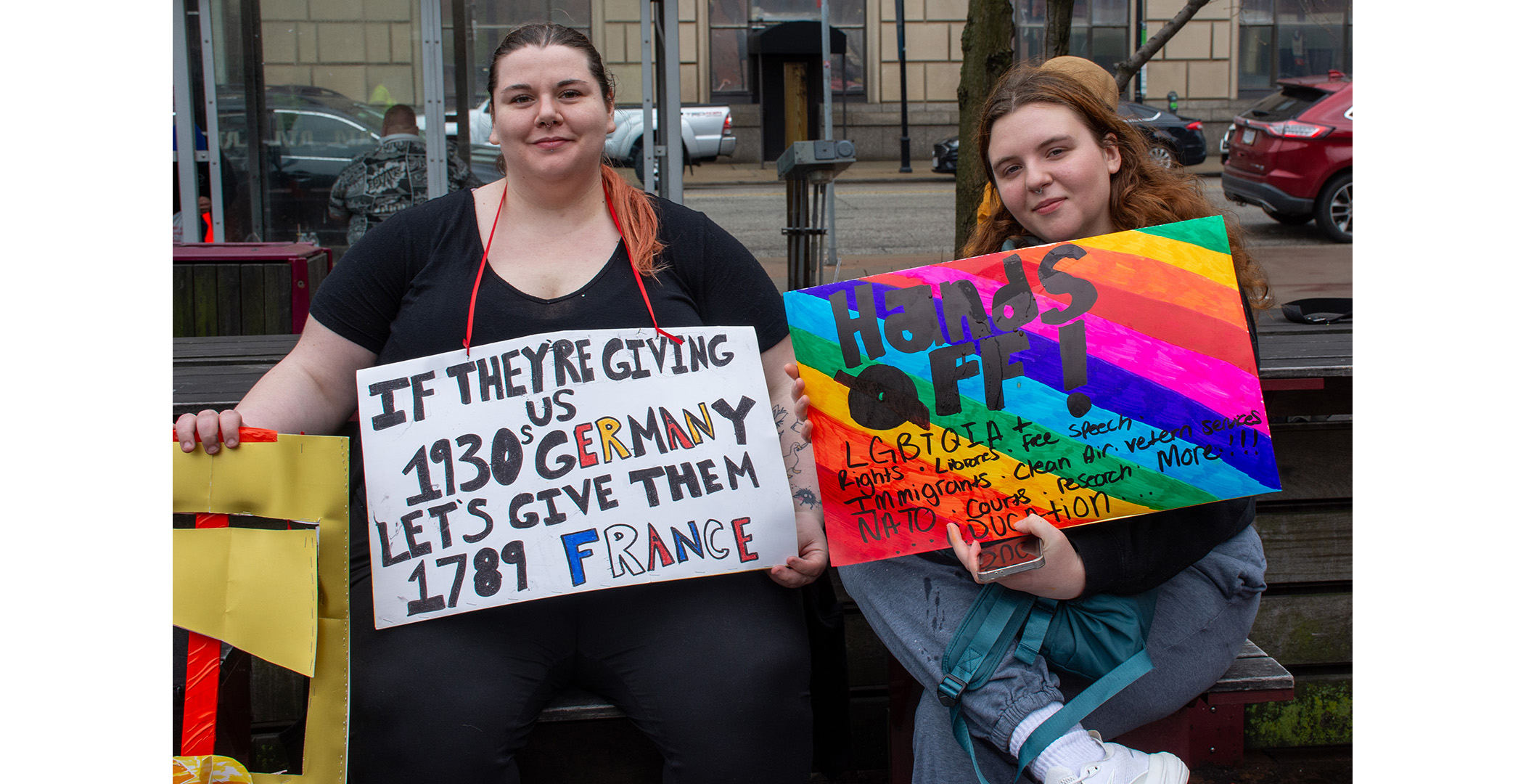 handsoff_protest6 Two women at the Cleveland Hands Off protest sitting next to each other. the woman on the left has a sign that says "If They're Giving Us 1930's Germany, Let's give them 1789 France. The woman on the right has a rainbow colorer sign saying "Hands Off LGBTQIA+ Rights - Libraries - Free Speech (sic) - Immigrants - Clean Air - Vetern (sic) Services - NATO - Courts - Research - Education - More!!!