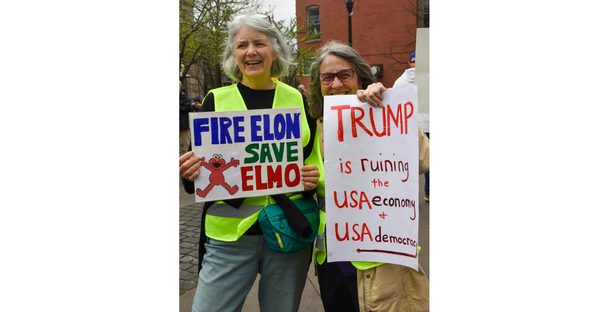 handsoff_protest7 Two woman holding signs; The sign on the left says "Fire Elon, Save Elmo" and the woman on the right holds one saying "Trump is ruing the USA economy & USA democracy"