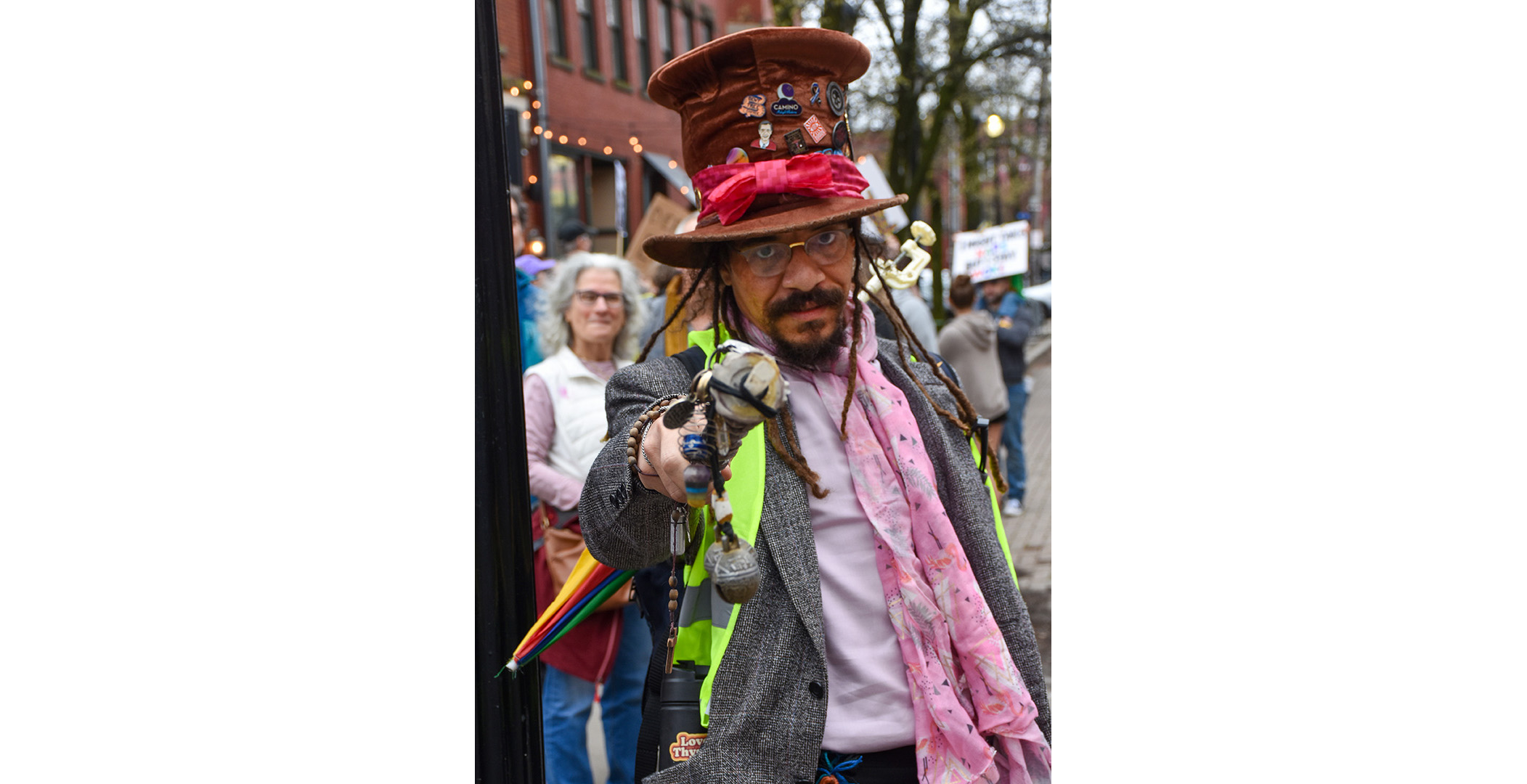 man_in_hat A man with a leather top hat filled with pins, thin dreadlocks. and a pin scarf. He is holding a multi-colored umbrella and point at the photographer with the hilt.