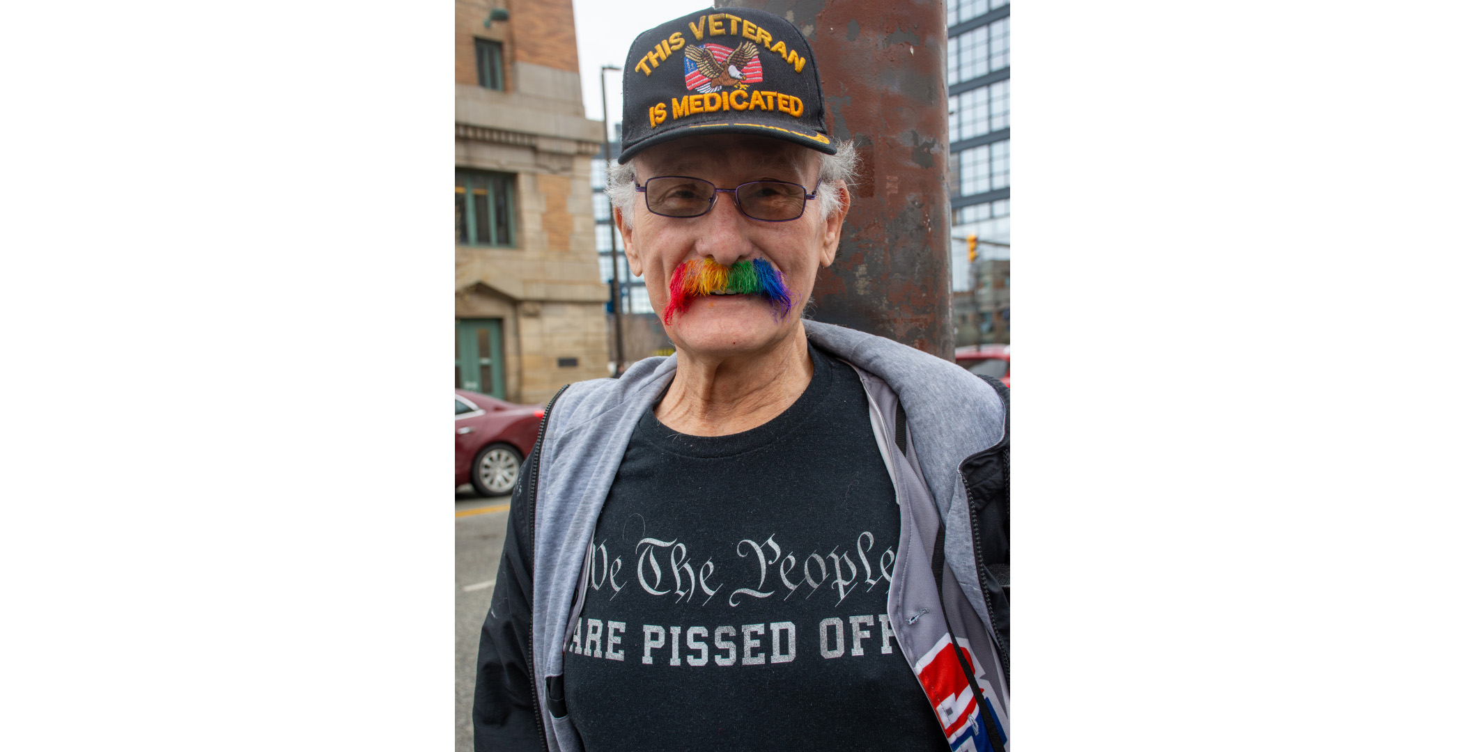rainbow moustache An older man with a rainbow colored moustache. His hat says "This veteran is Medicated with an eagle and American flag. His shirt says "We the people are pissed off"
