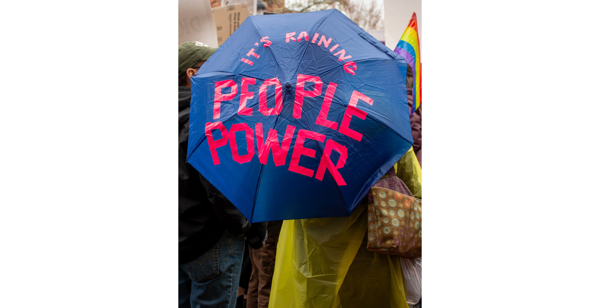 raining people power Back of woman in yellow rain slicker holding a blue umbrella. In read, the umbrella says "It's raining People Power"